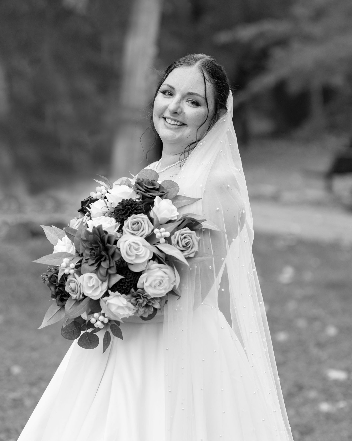 Black and white bridal portrait of smiling bride in wedding gown and veil holding large floral bouquet outdoors.