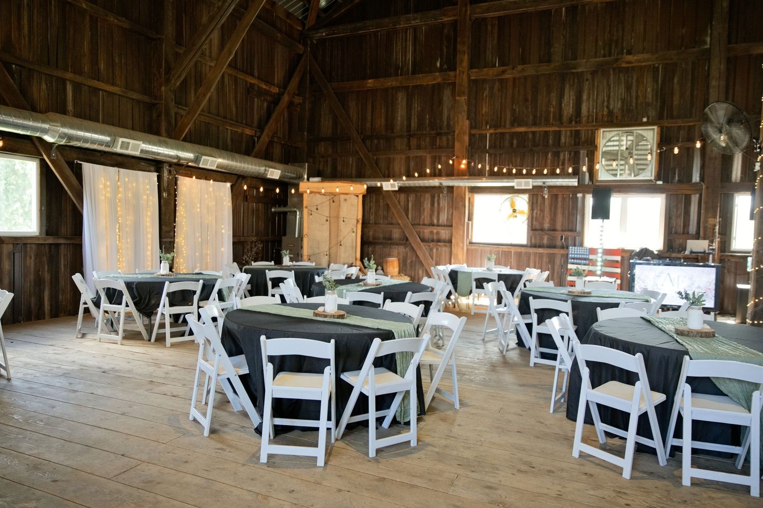 Rustic barn wedding reception venue with white folding chairs, black draped tables, string lights, and wooden walls.
