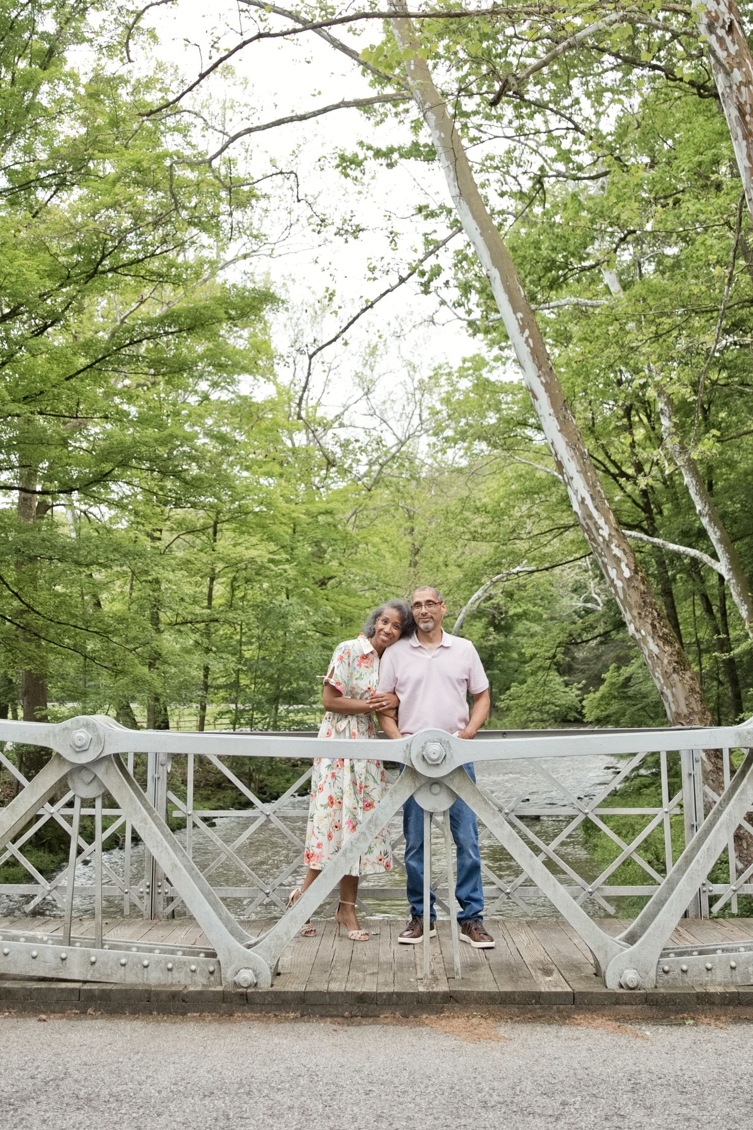 Couple posing on a white bridge surrounded by lush green trees in a park setting.