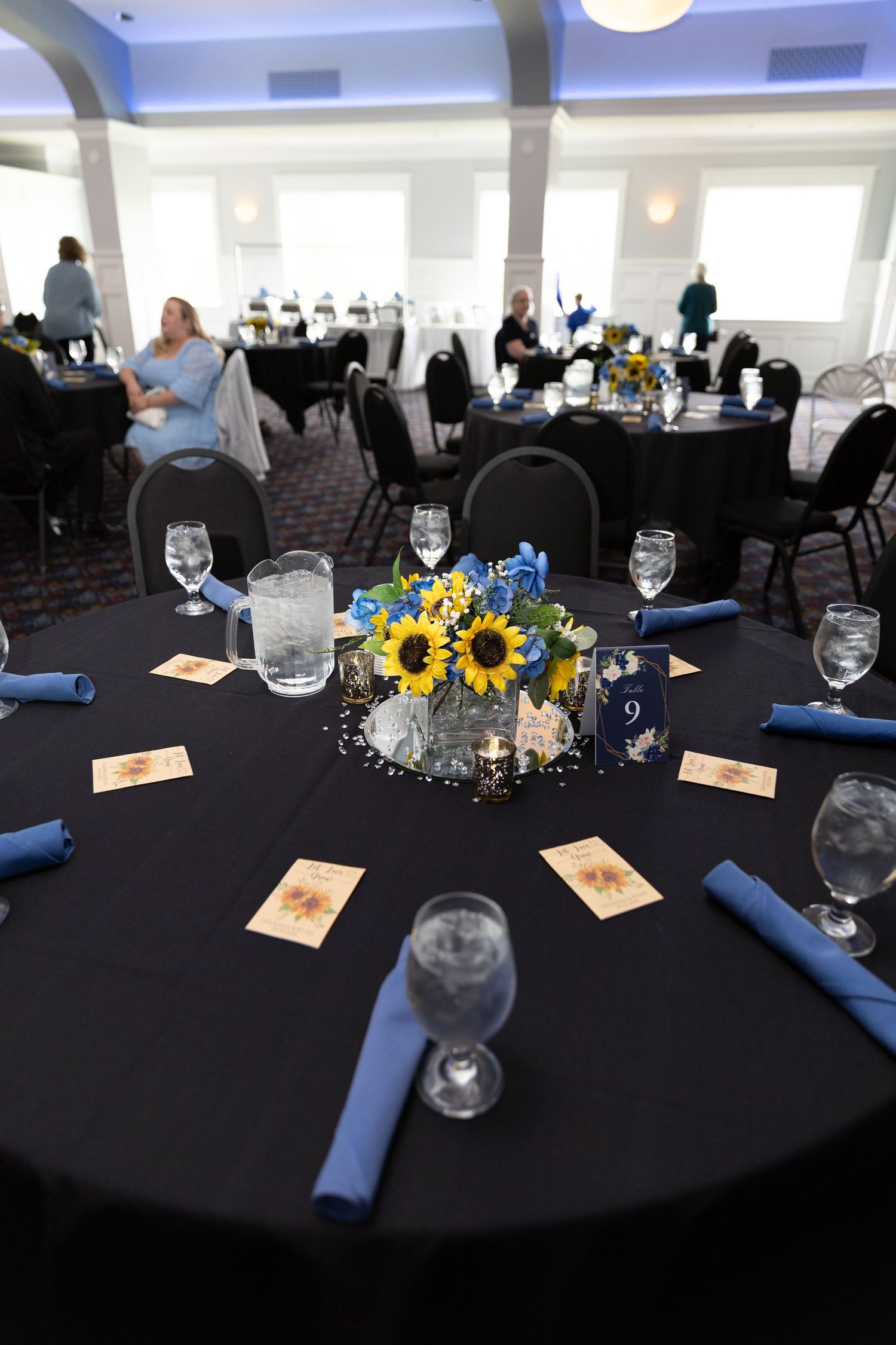 Elegant banquet hall with black tables, blue napkins, and sunflower centerpieces set for a formal event.