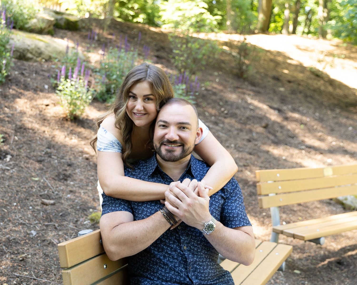 Happy couple posing on a park bench, woman hugging man from behind, smiling outdoors in a lush garden setting.