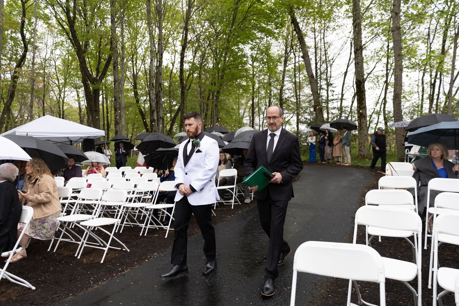 Two men in formal attire walk down an outdoor wedding aisle on a rainy day surrounded by guests with umbrellas.