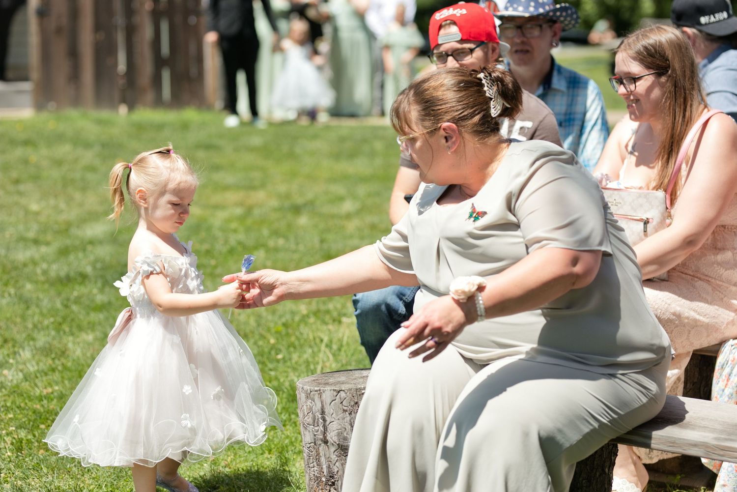 Young flower girl in white dress handing a flower to a seated woman in sage green dress at outdoor wedding.