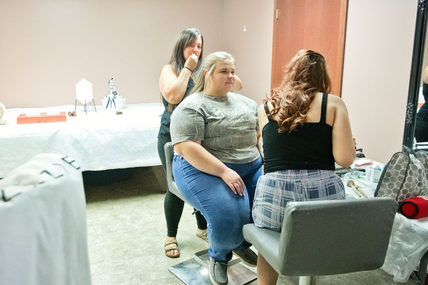 Three women in a beauty salon room, one receiving makeup application while others observe nearby.
