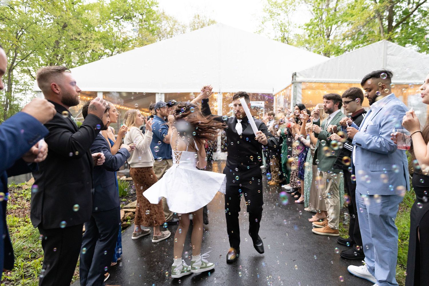 Bride and groom exit wedding reception under bubbles, surrounded by cheering guests near a white tent.