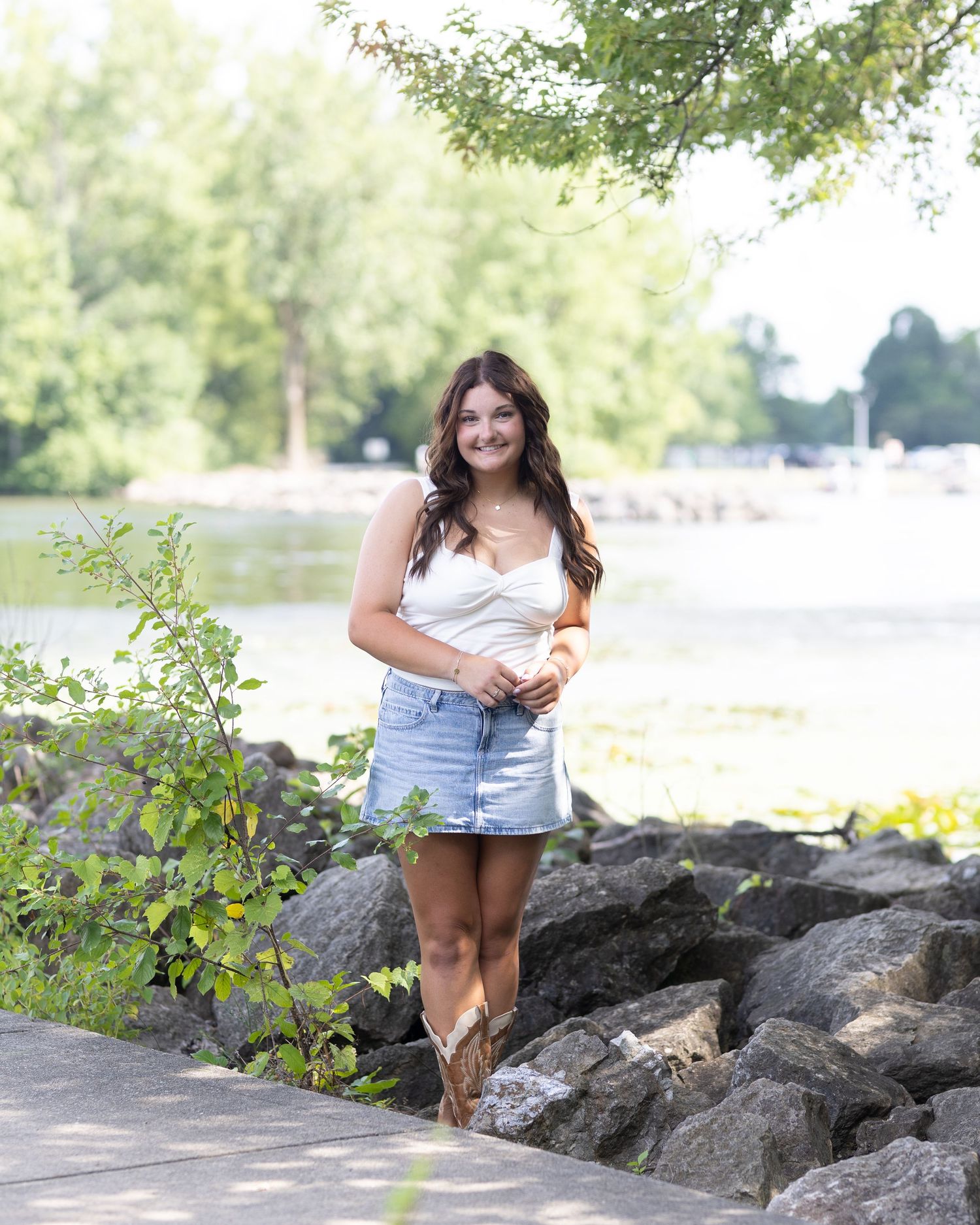 Summer portrait by the water with cowboy boots, denim shorts and a white top against a rocky shoreline.
