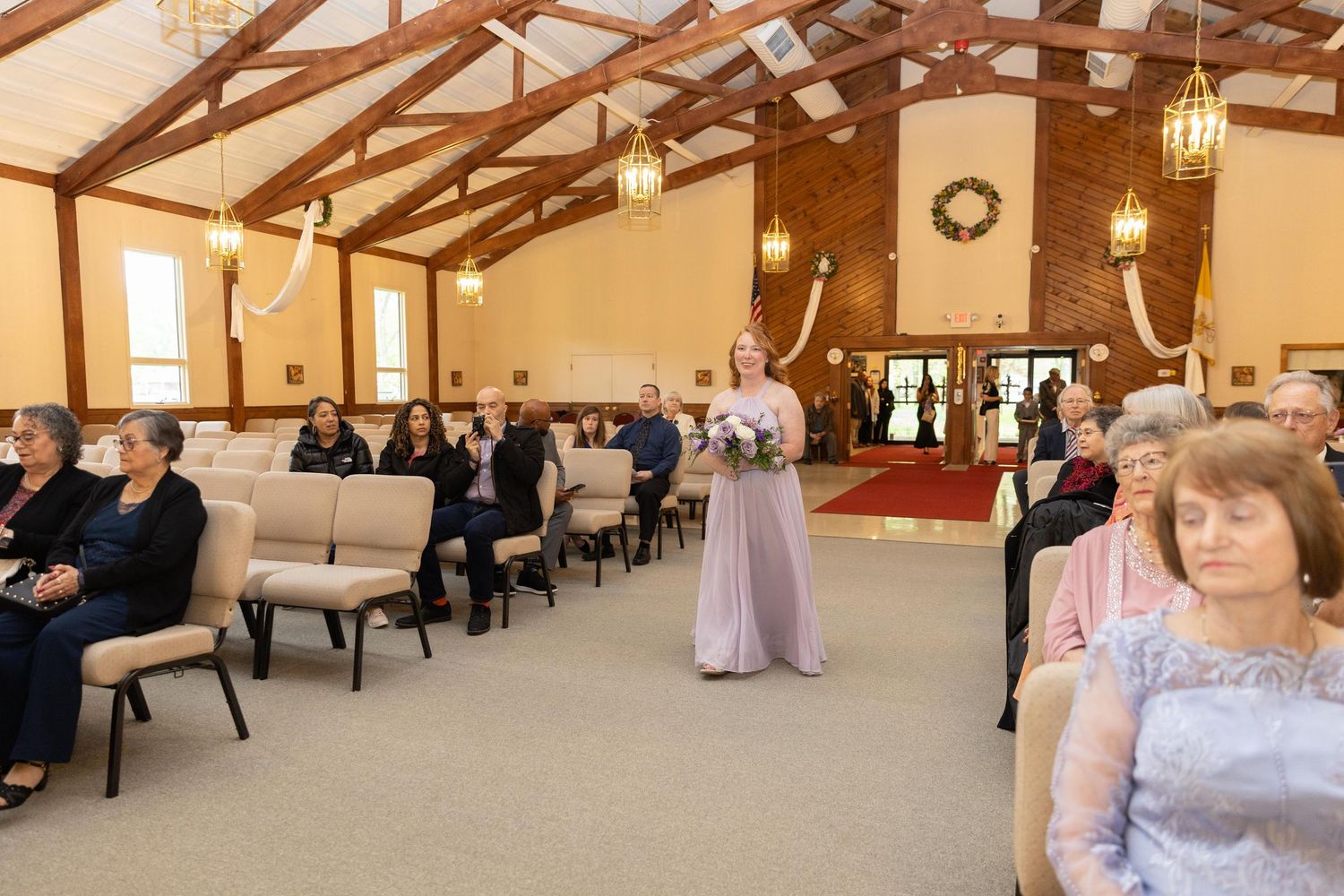 Bridesmaid in lavender gown walks down the aisle of an elegant chapel during a wedding ceremony.
