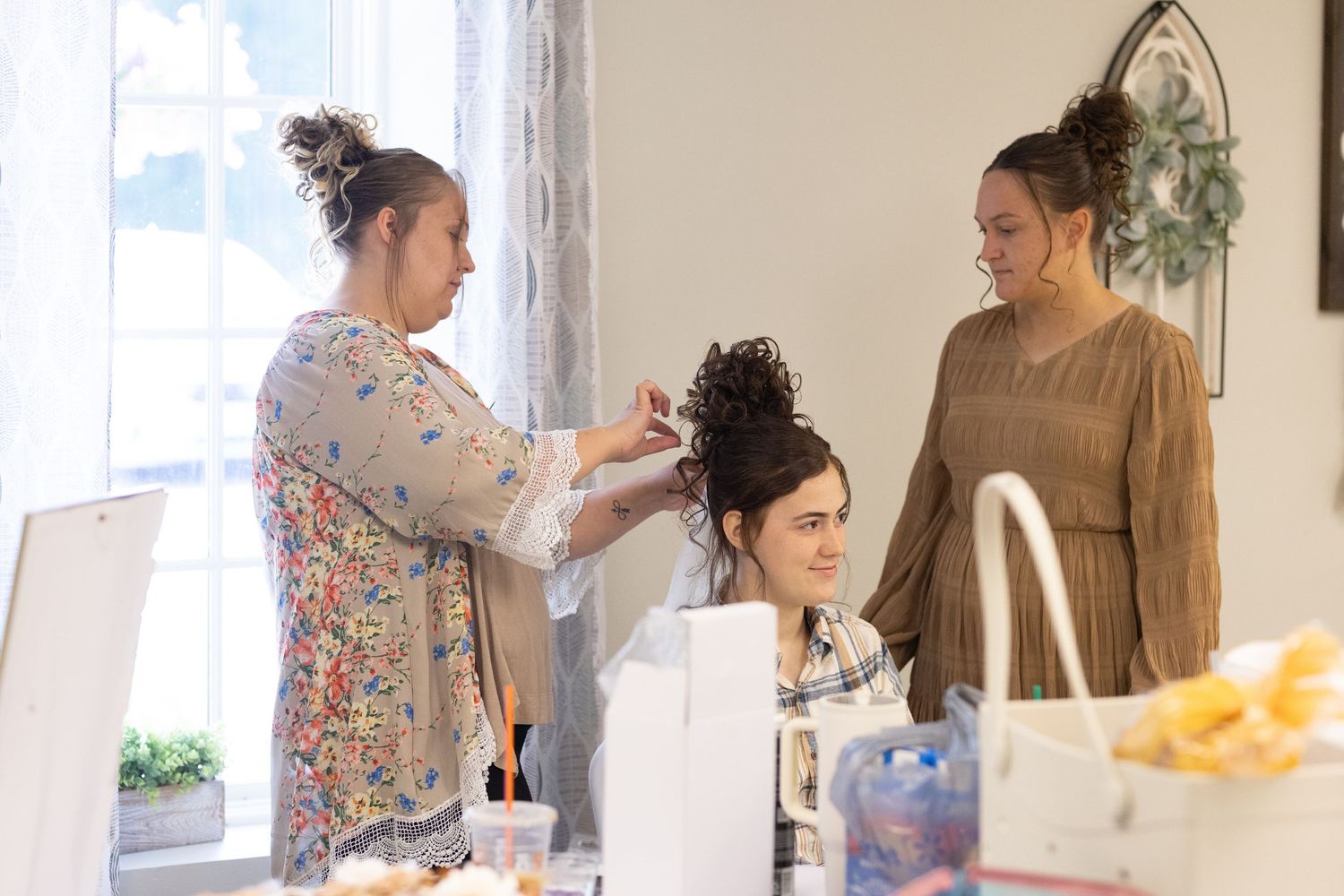 Three people prepare for a special event with one having their hair styled in a bright room.