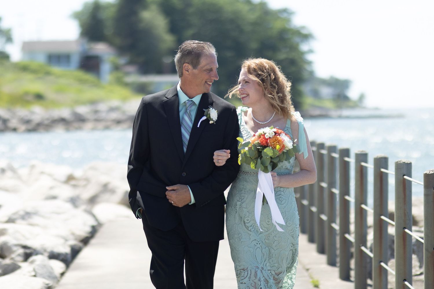 A couple walks along a seaside boardwalk during a wedding celebration with colorful bouquet.