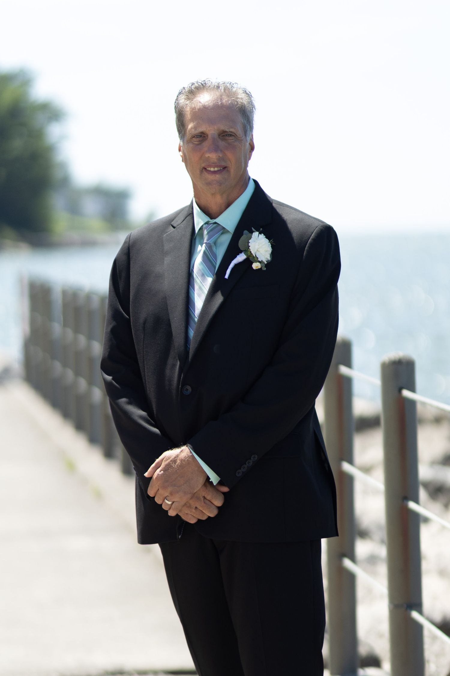 A person in a black suit and tie stands by a wooden railing near the ocean.