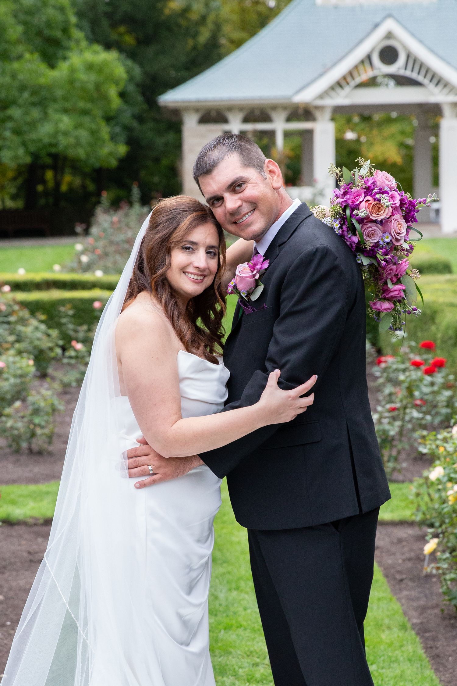 Wedding couple embraces in front of white gazebo surrounded by blooming roses.