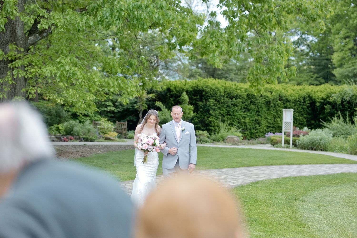 Bride and groom walking down outdoor garden aisle on sunny wedding day with lush green trees and hedges.