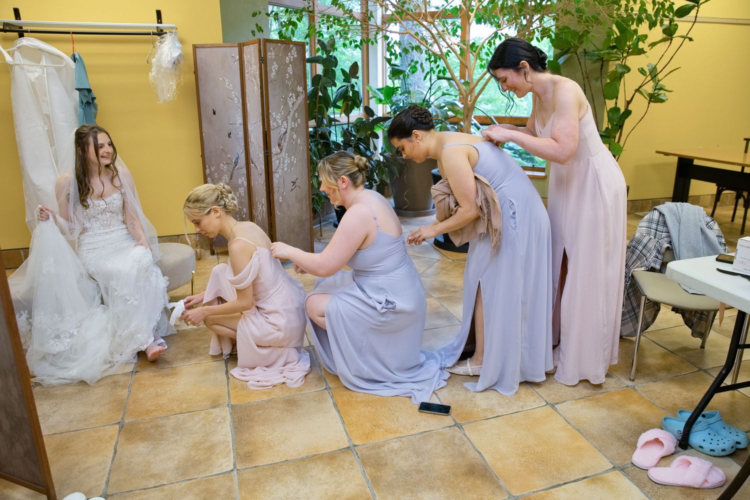 Bridesmaids in lavender gowns help bride prepare in a bright dressing room with a white wedding dress hanging nearby.