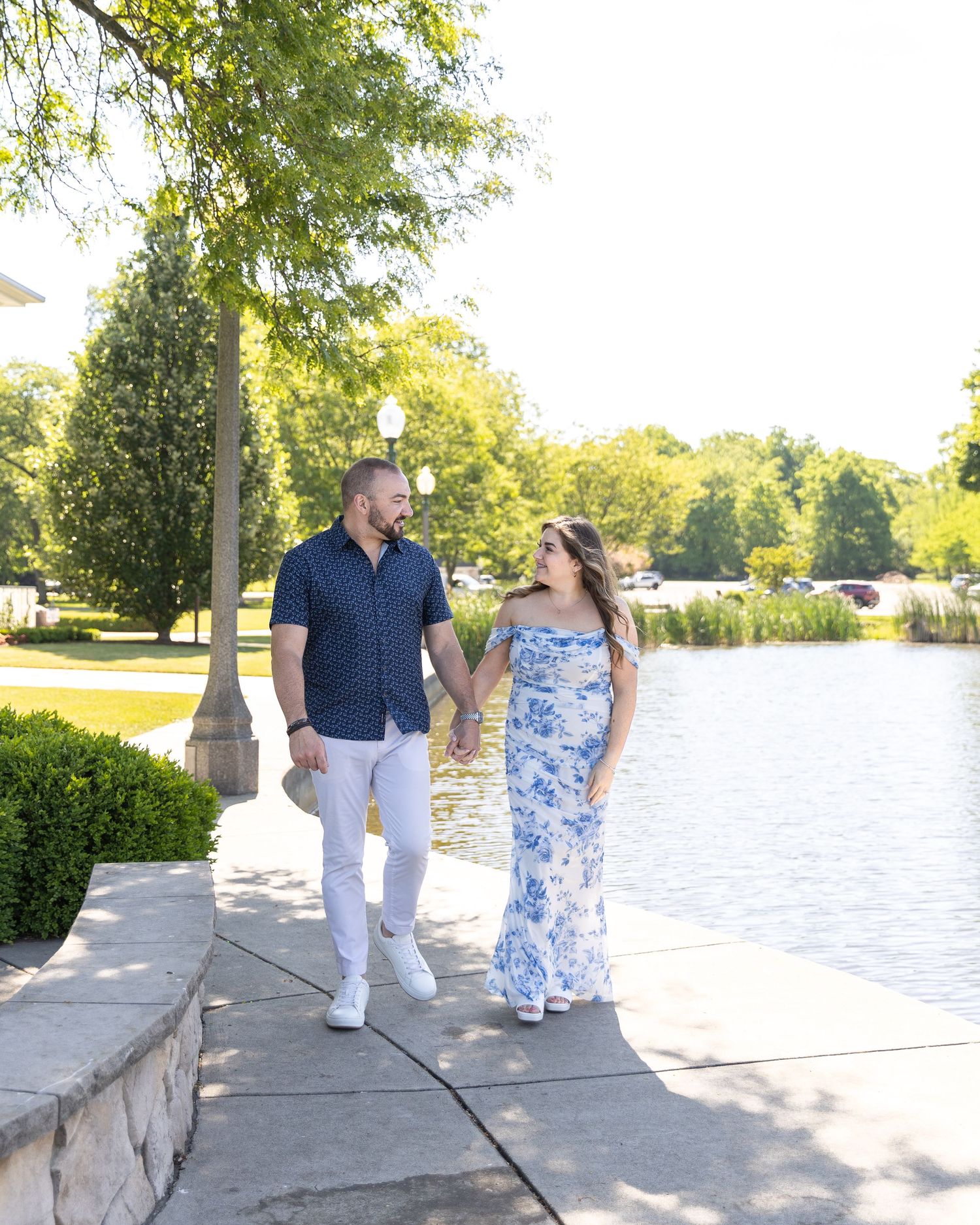 Happy couple walking hand in hand along a scenic waterfront path surrounded by lush green trees.
