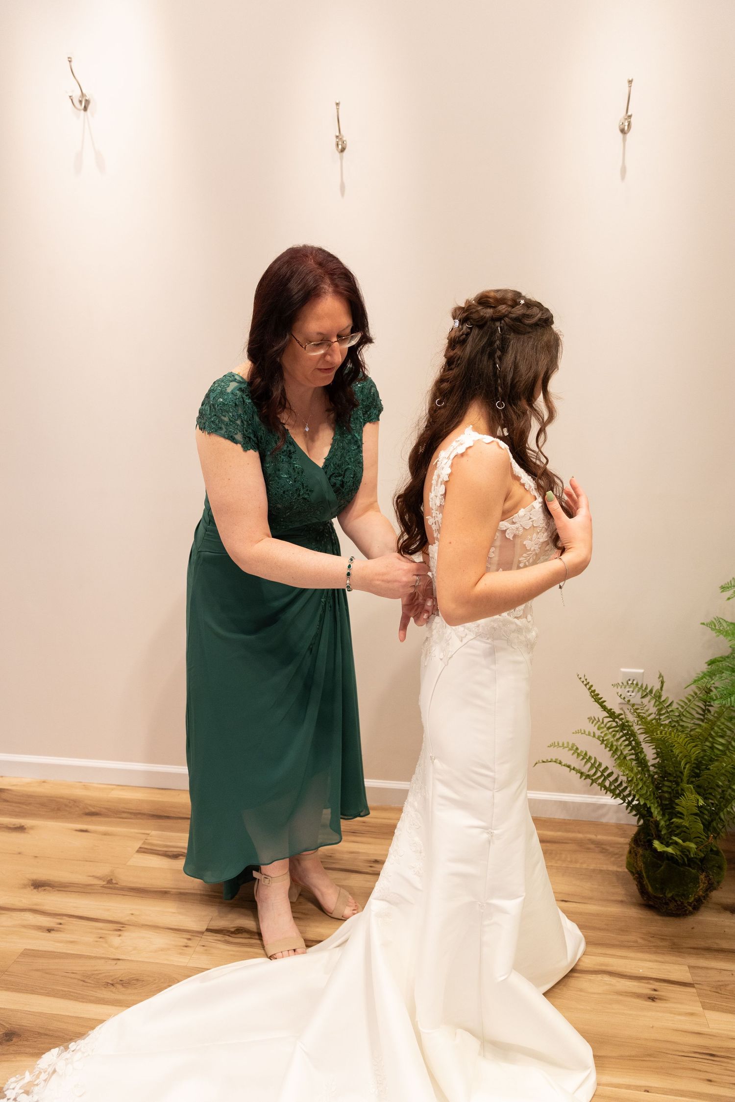 A bride in a white gown with a train gets help from a woman in a green dress in a warmly lit room.
