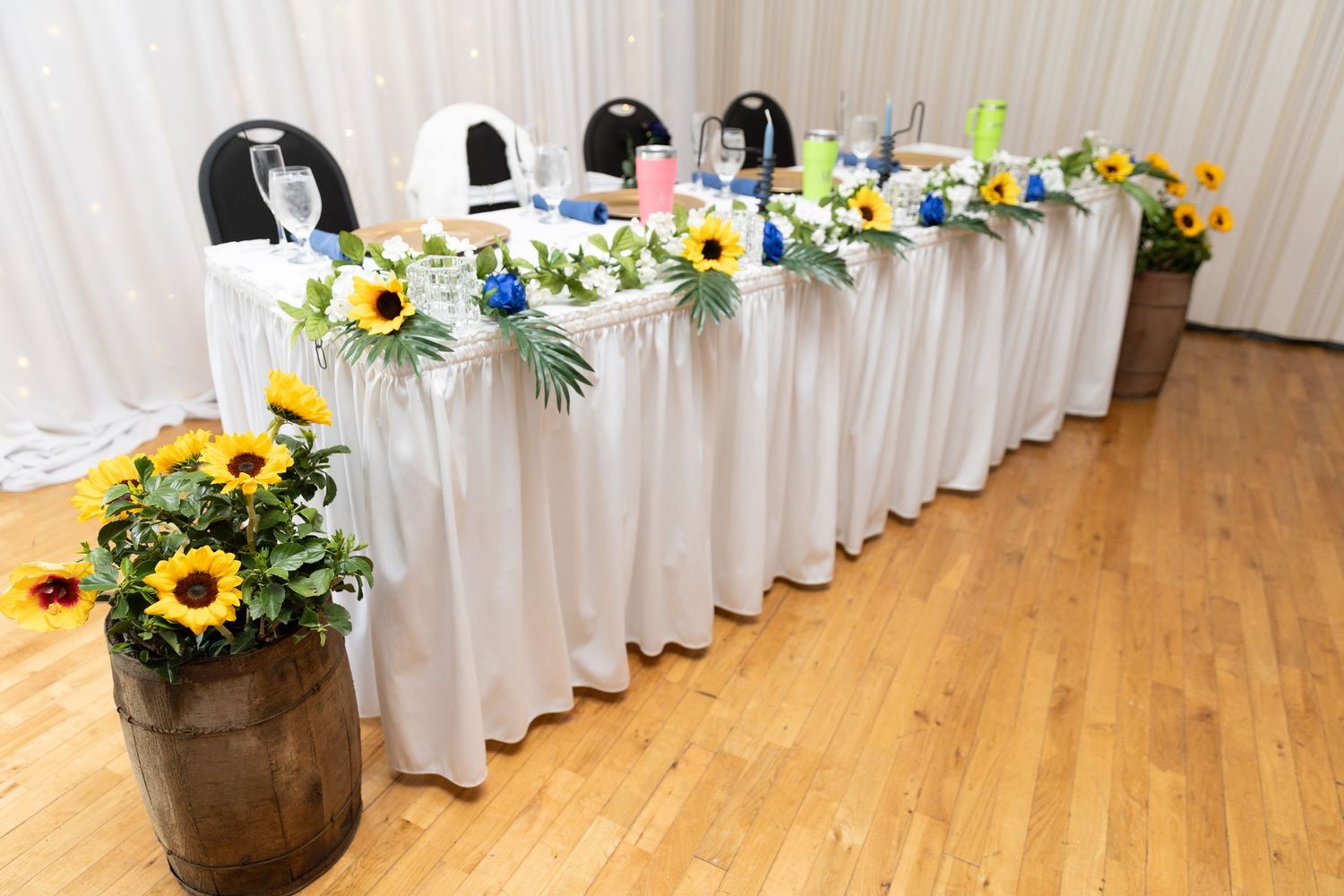 Elegant wedding head table with white skirting, sunflower arrangements, and rustic barrel centerpieces on a wooden floor.