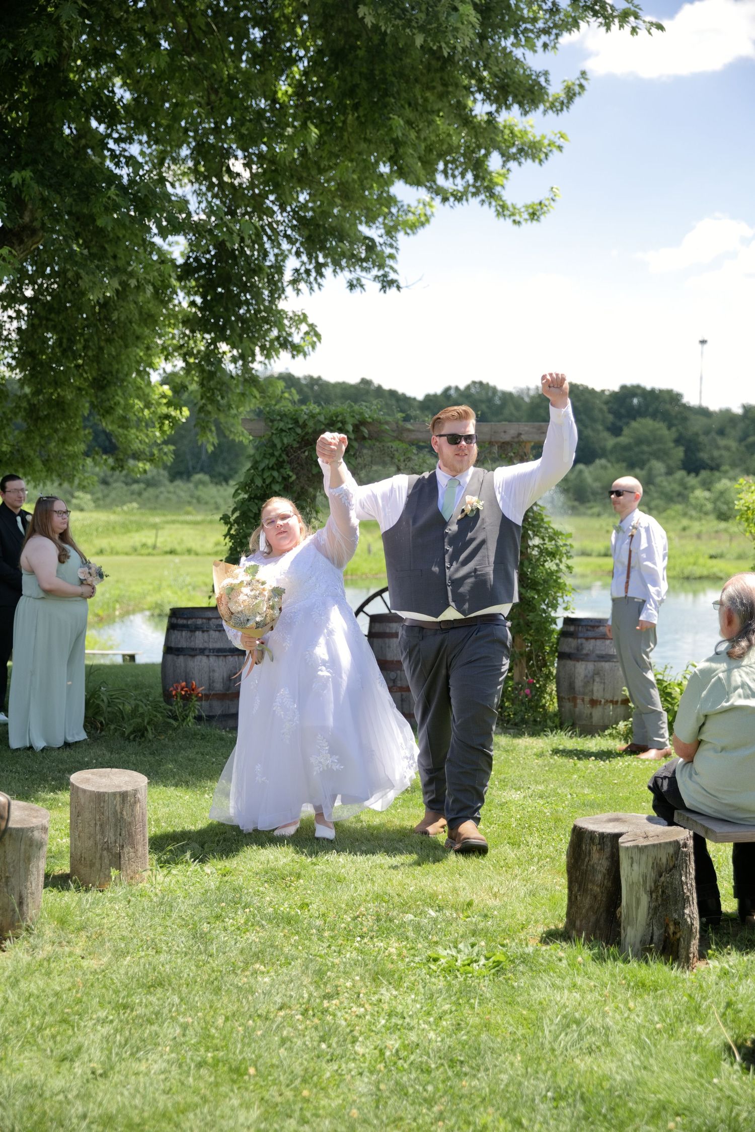 Joyful newlyweds raise their arms in celebration at an outdoor wedding ceremony on a sunny green lawn.