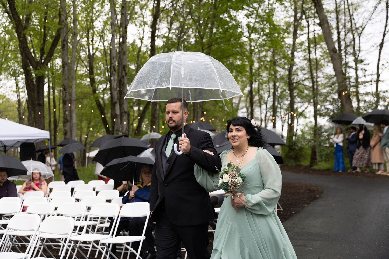 Bride in green gown and groom in black tuxedo walk under clear umbrella at outdoor rainy wedding ceremony.