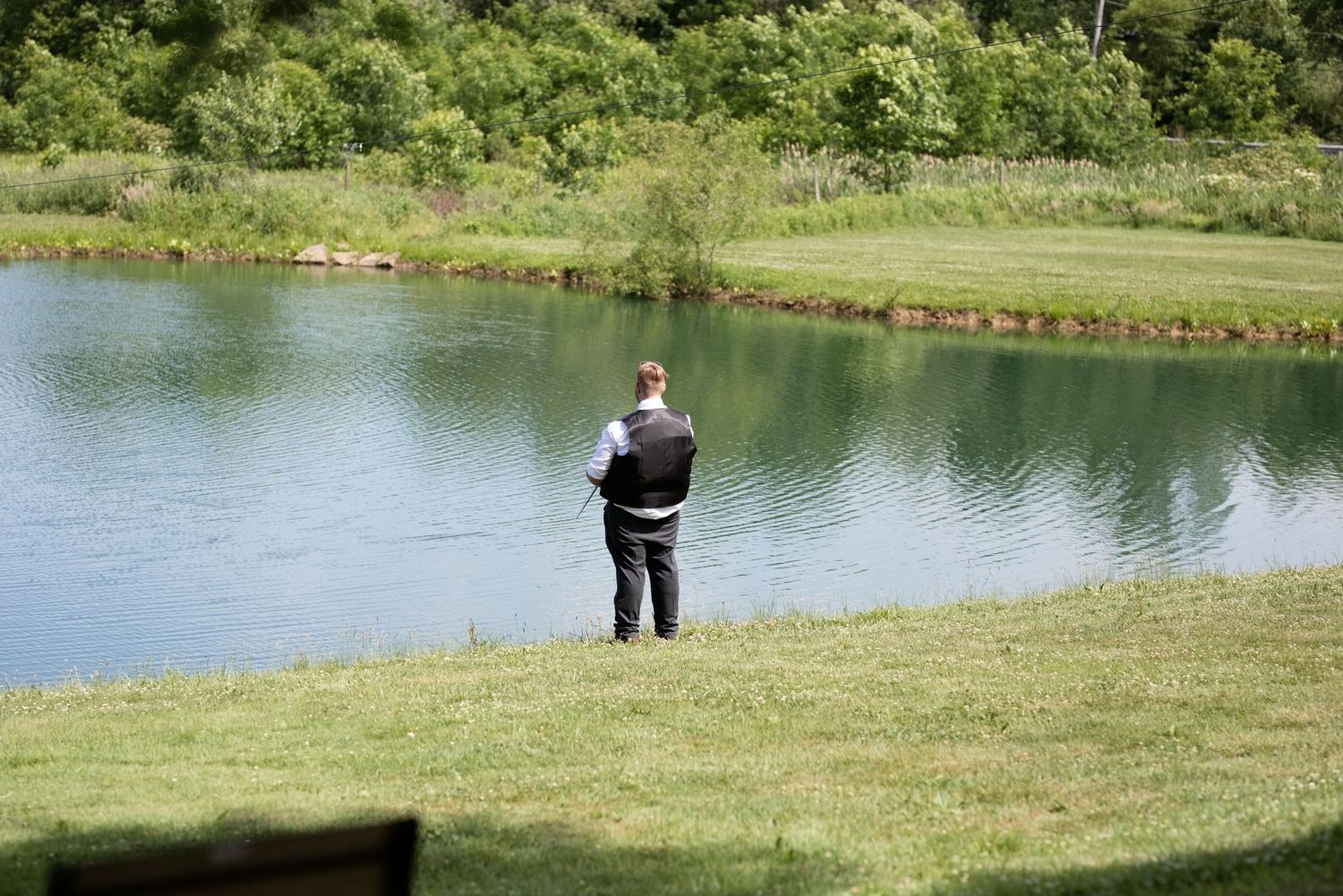 Man fishing alone by a serene pond surrounded by lush green trees and grass.