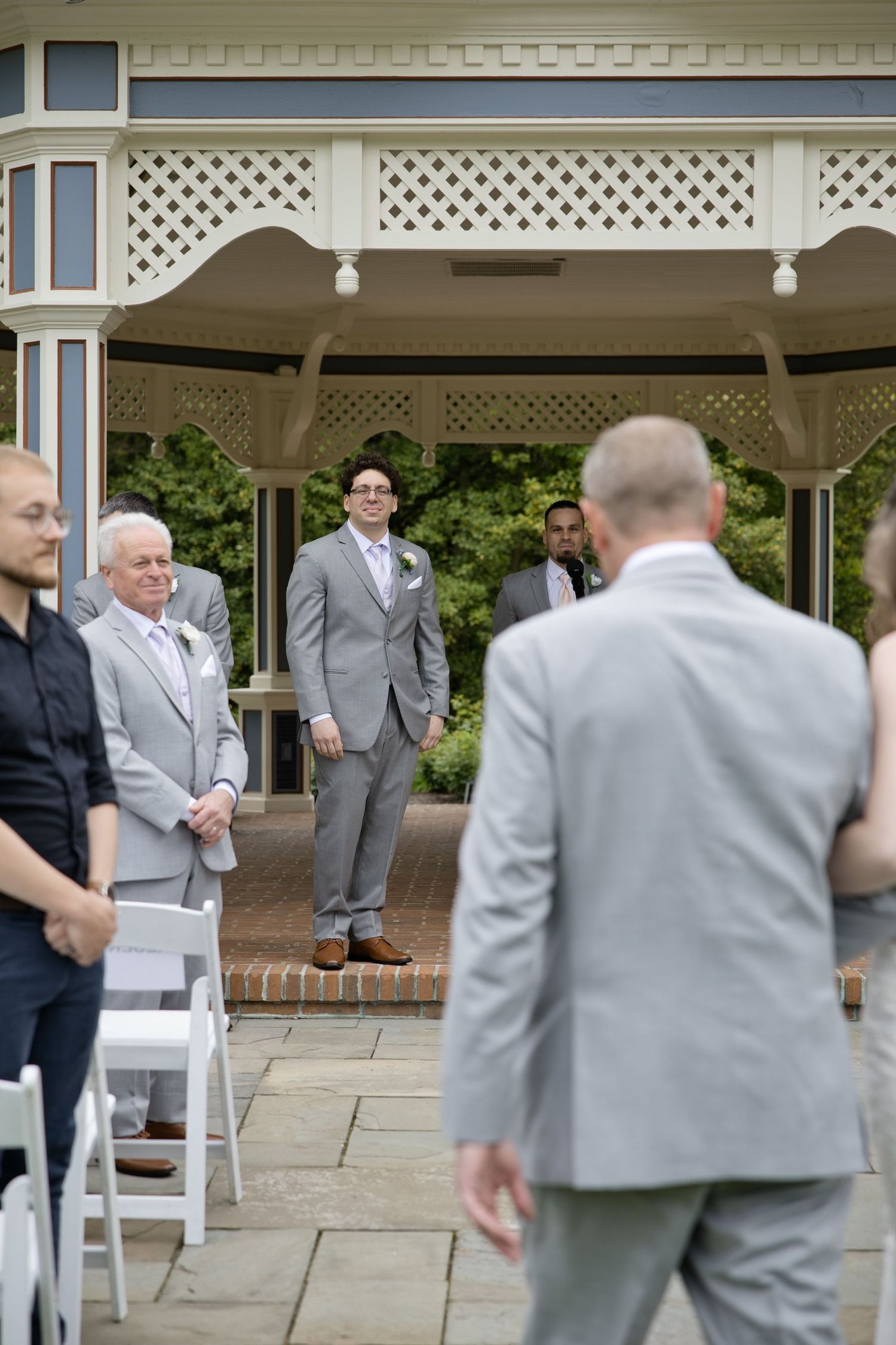 Groom in gray suit stands at gazebo altar awaiting bride during outdoor wedding ceremony.
