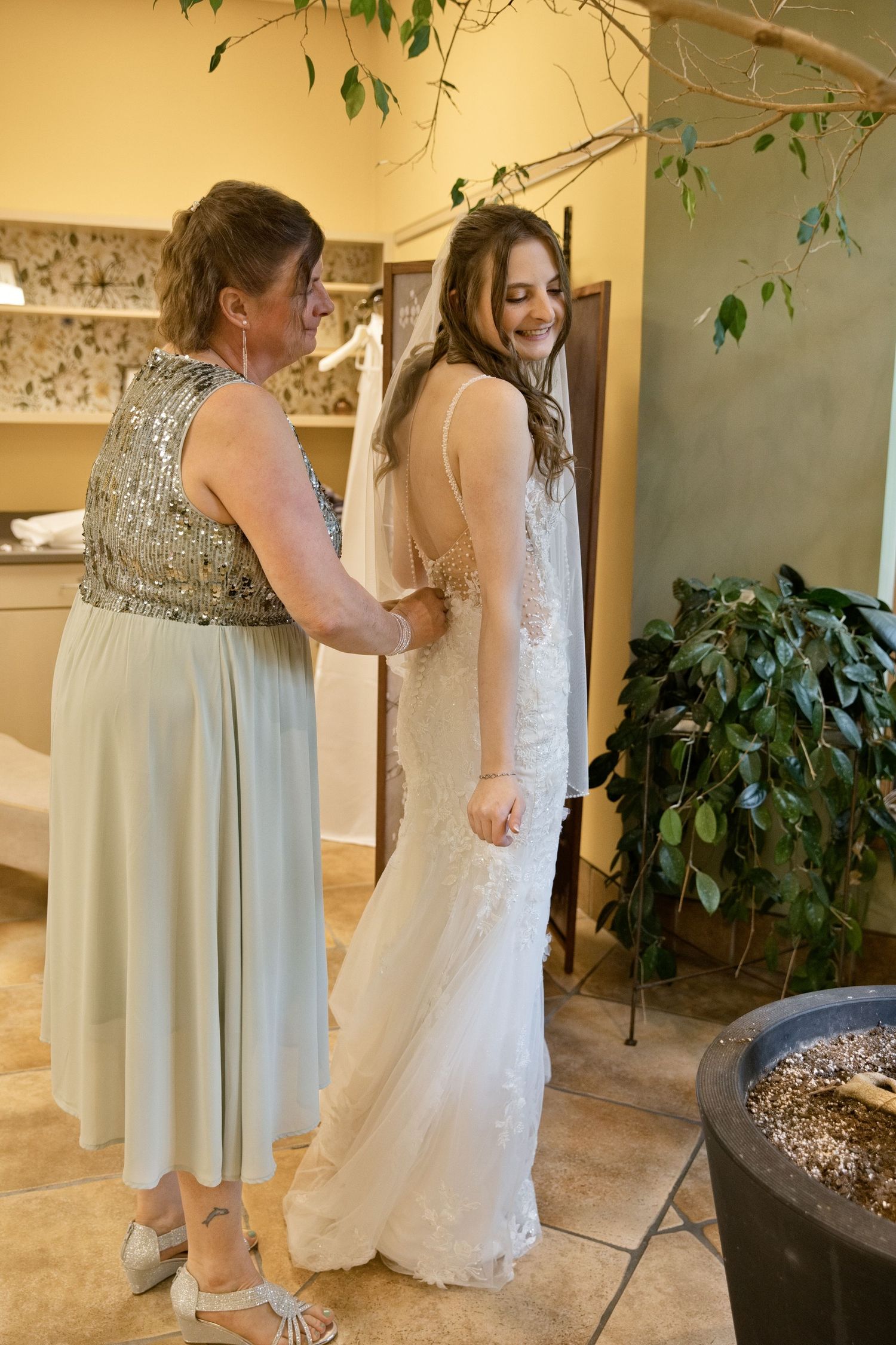 A bride in a white lace gown smiles as an older woman in a mint sequin dress helps fasten her wedding dress.