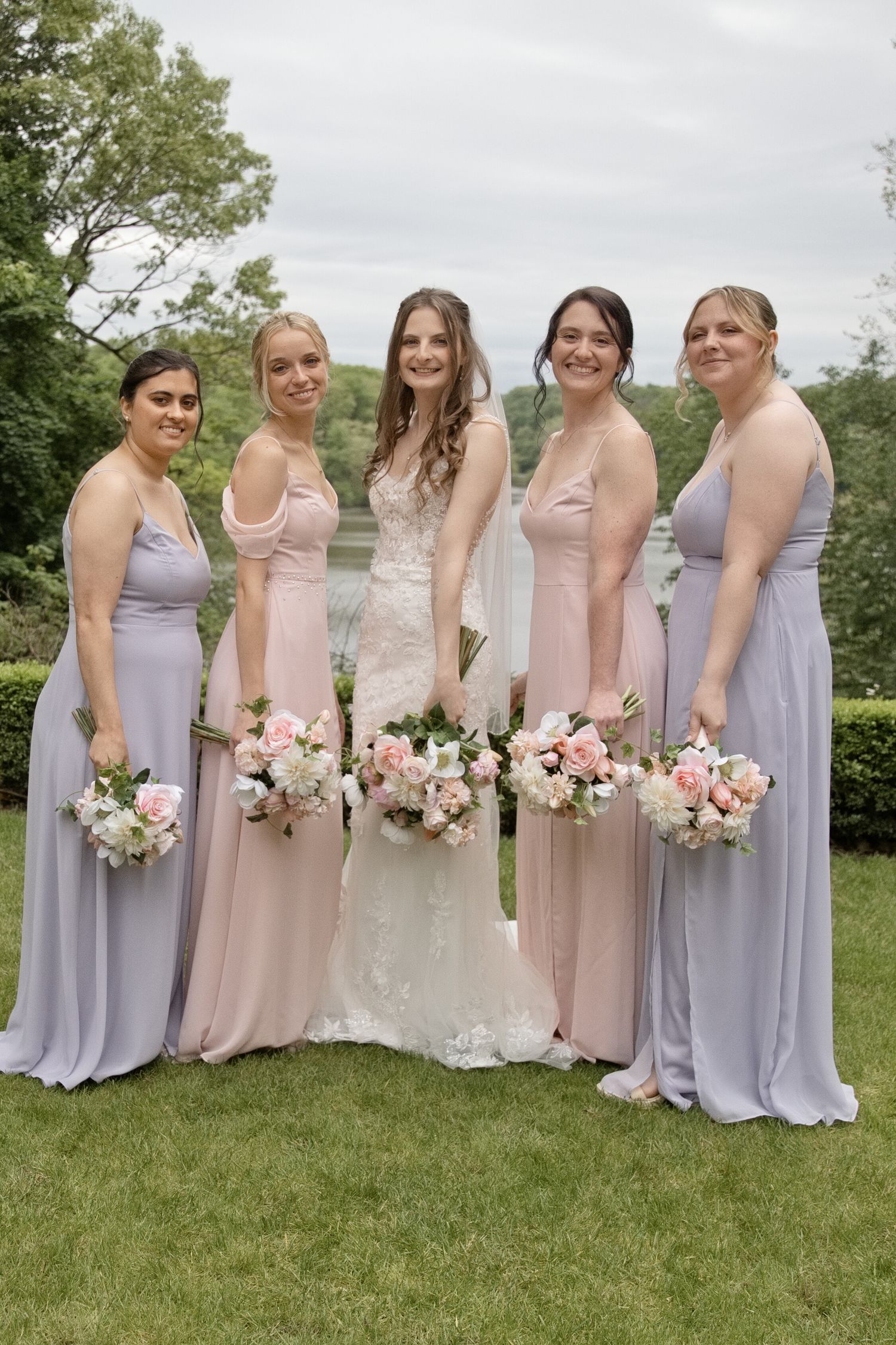 Bride surrounded by four smiling bridesmaids in pink and lavender gowns holding bouquets outdoors.