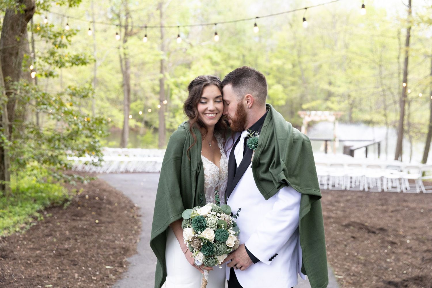 Bride and groom share a romantic moment outdoors, wrapped in a green cape, surrounded by misty trees and string lights.