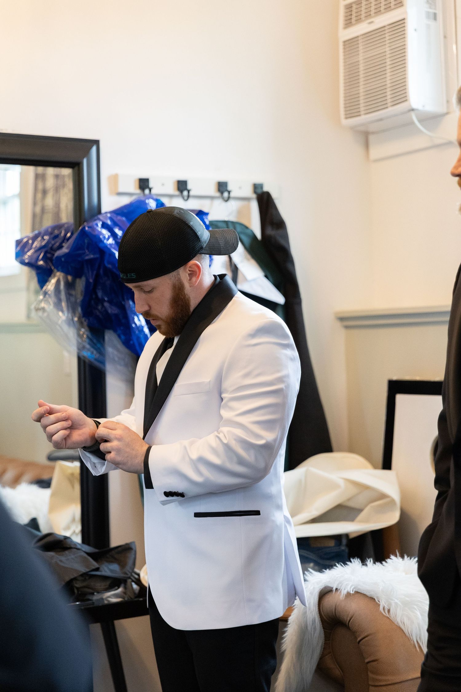 Man in white tuxedo jacket and black cap adjusting cufflinks in a dressing room before a formal event.