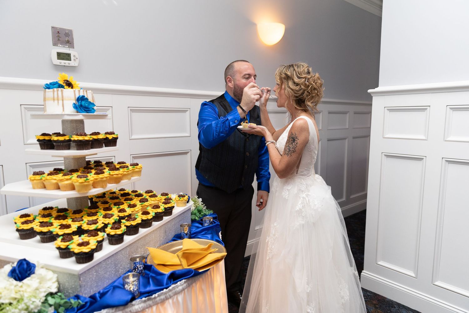 Bride and groom sharing wedding cake at reception with sunflower cupcake tower display in elegant white venue.
