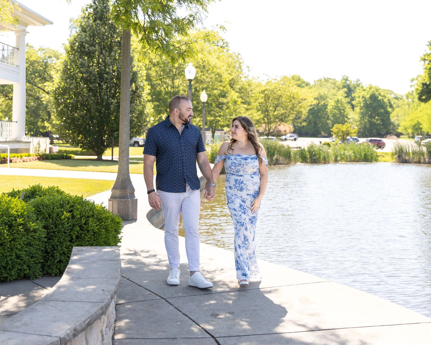 Couple holding hands and walking along a scenic waterfront path surrounded by lush green trees on a sunny day.