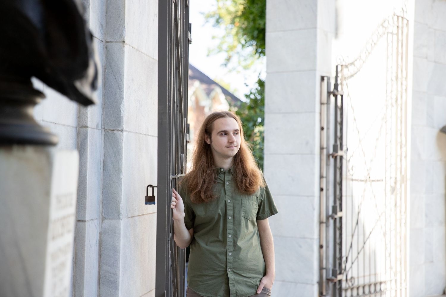 A person in olive green clothing stands between white pillars in an architectural setting.