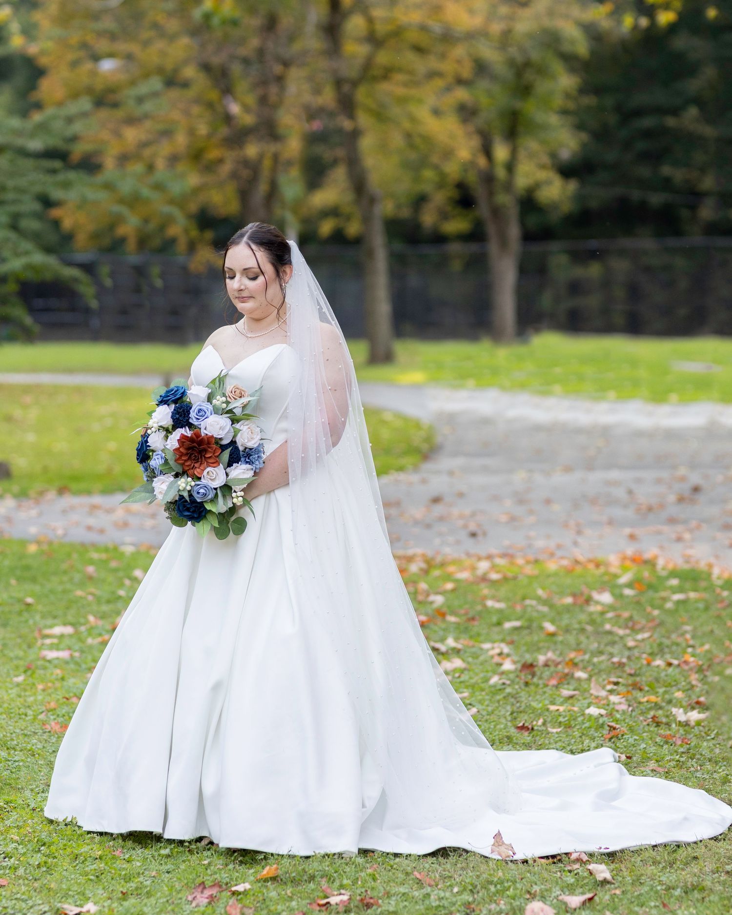 A bride in a flowing white wedding gown and veil poses outdoors with a blue and burgundy bouquet in autumn.