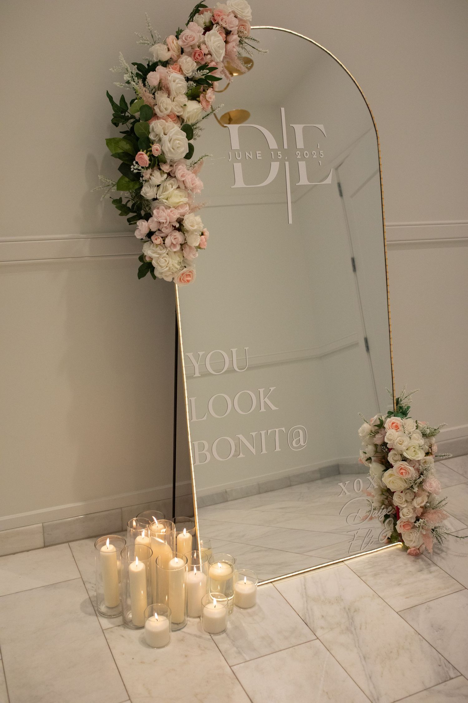 Elegant arched mirror with pink floral decorations and candles displaying 'You Look Bonita' text on marble floor.