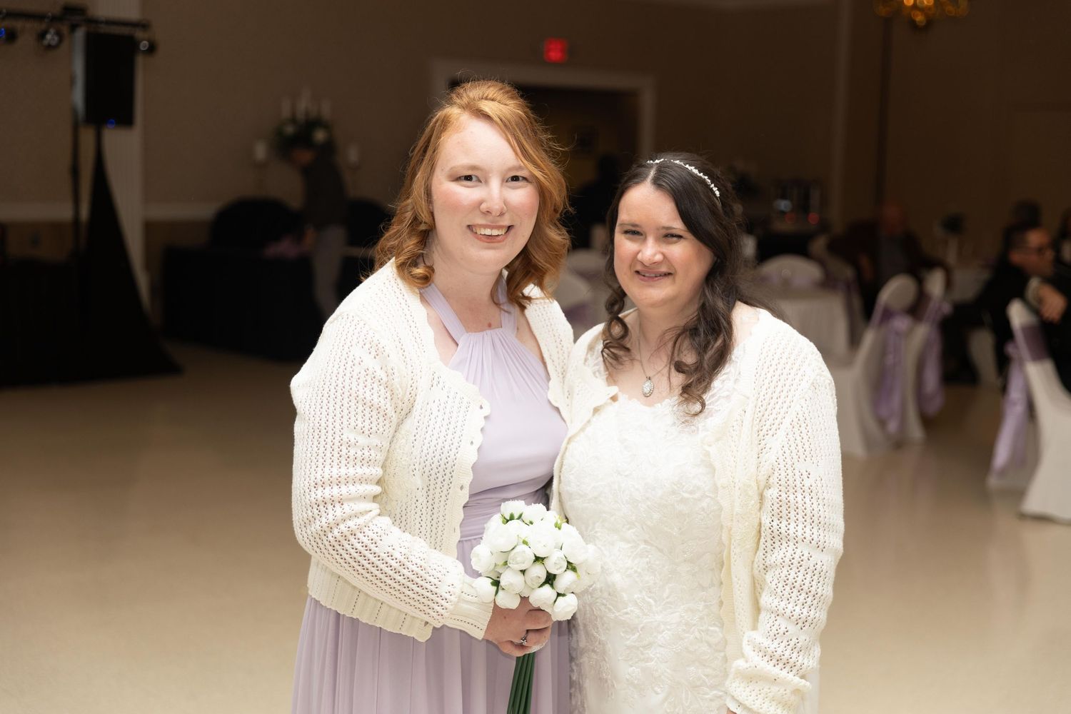 Two women in white cardigans pose together at a wedding reception, one in a bridal gown holding a bouquet.
