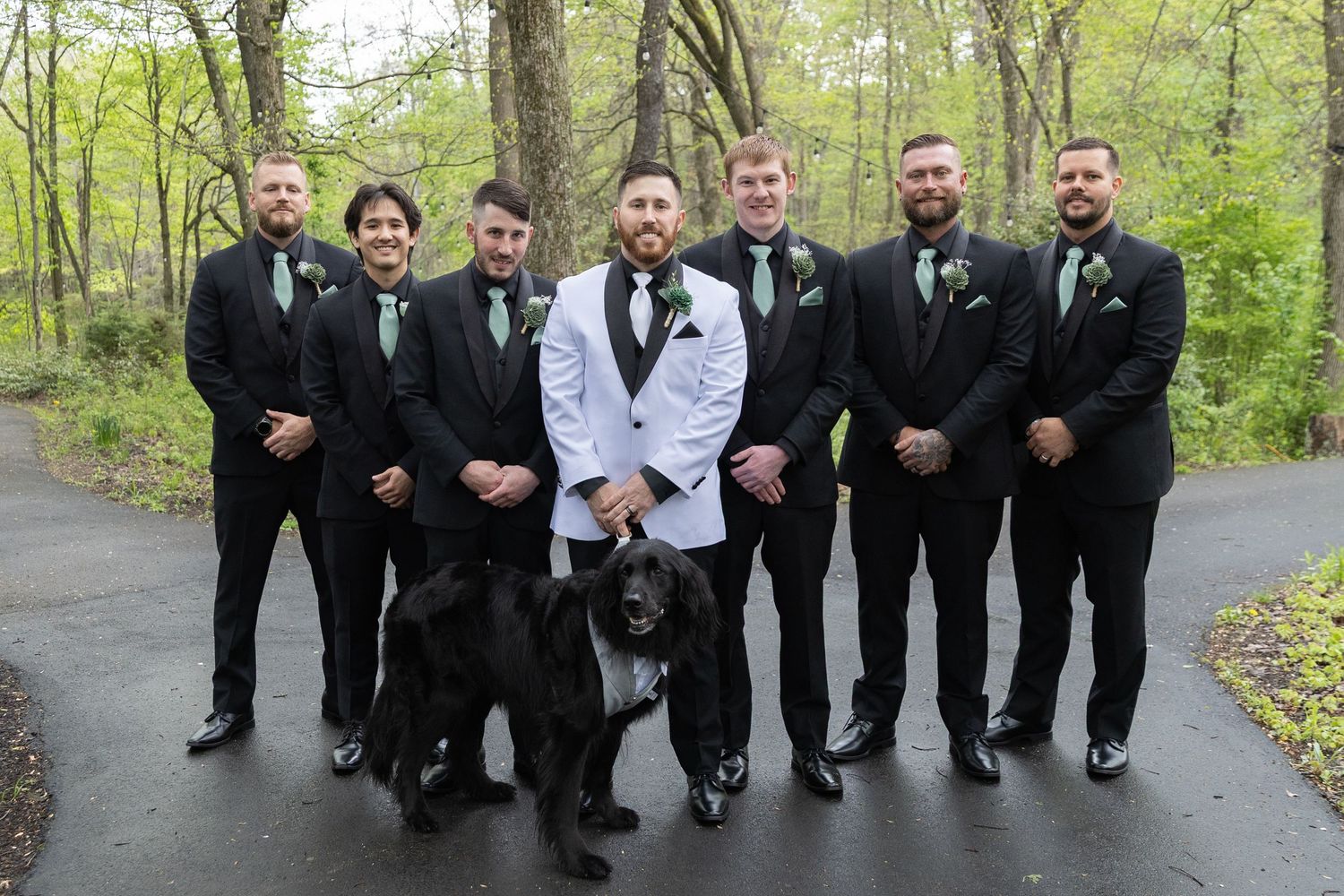 Groom in white tuxedo with groomsmen in black suits and green ties posing with a black dog outdoors.