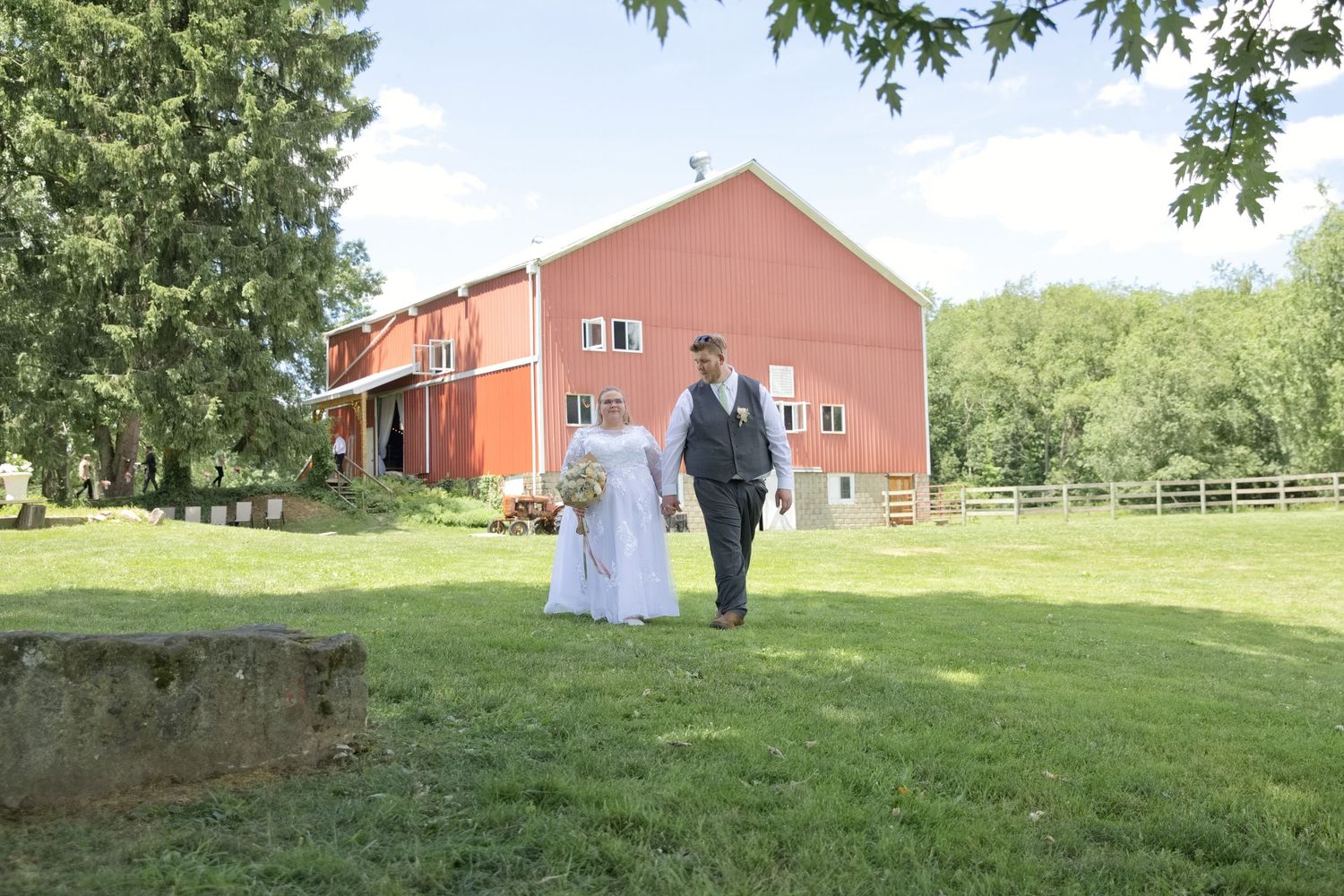 Bride and groom walking hand in hand on green lawn with red barn backdrop on sunny wedding day.