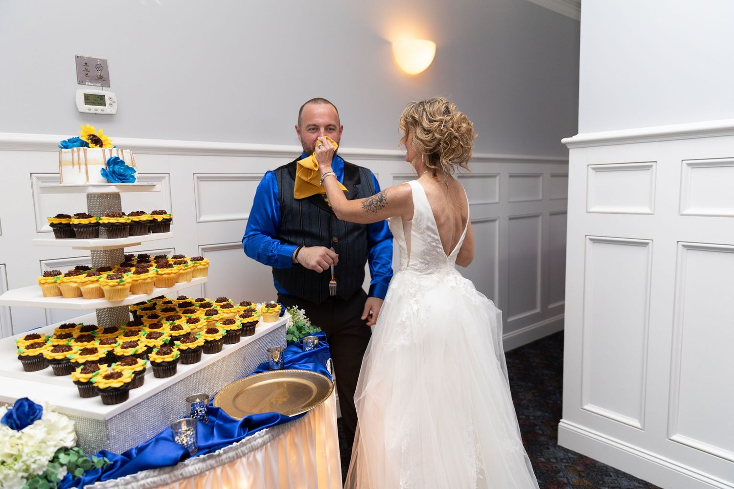Bride feeding groom cake at wedding reception with cupcake tower display in blue and gold decorations.