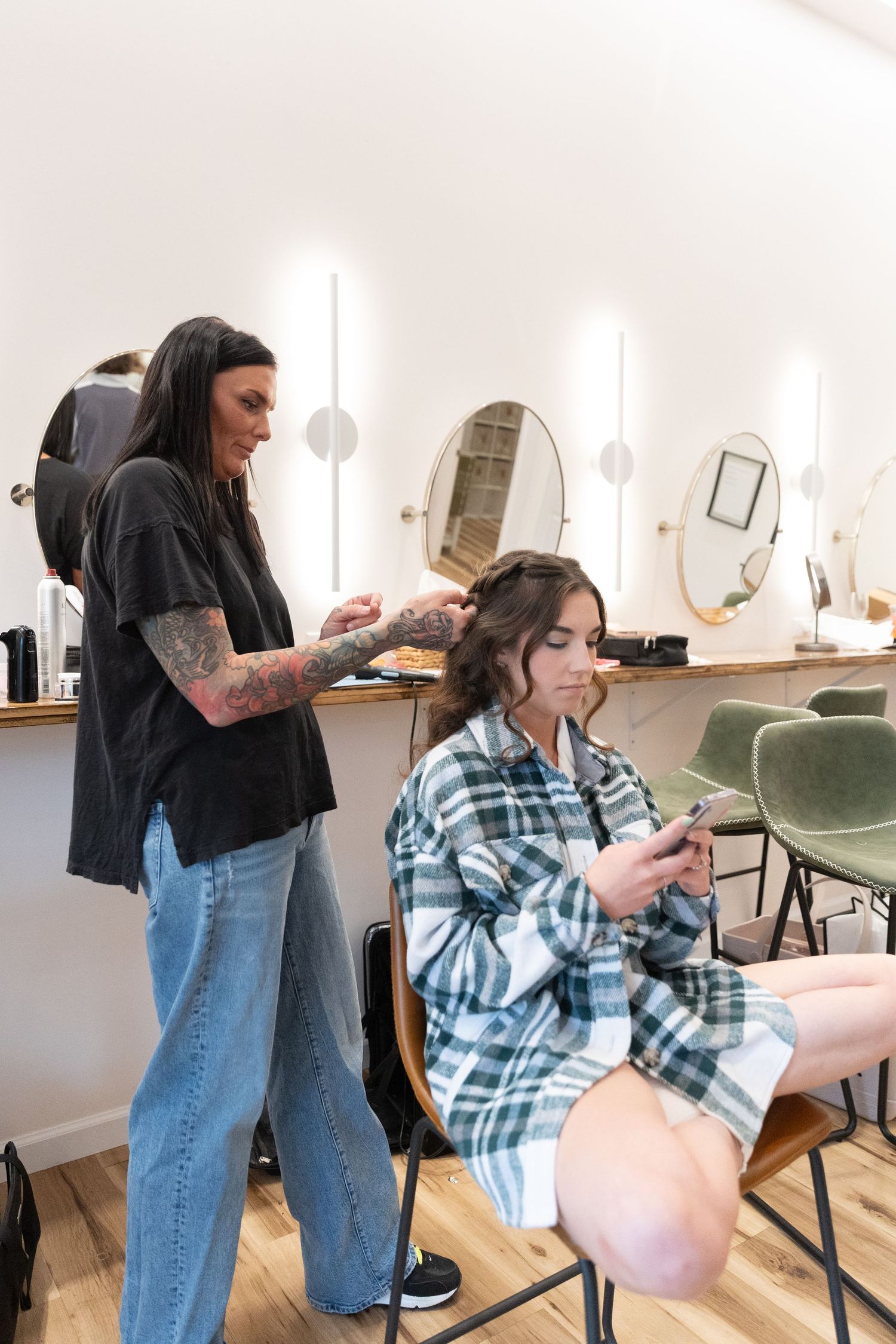 Tattooed hairstylist styling brunette client's hair in modern salon with round mirrors and warm lighting.