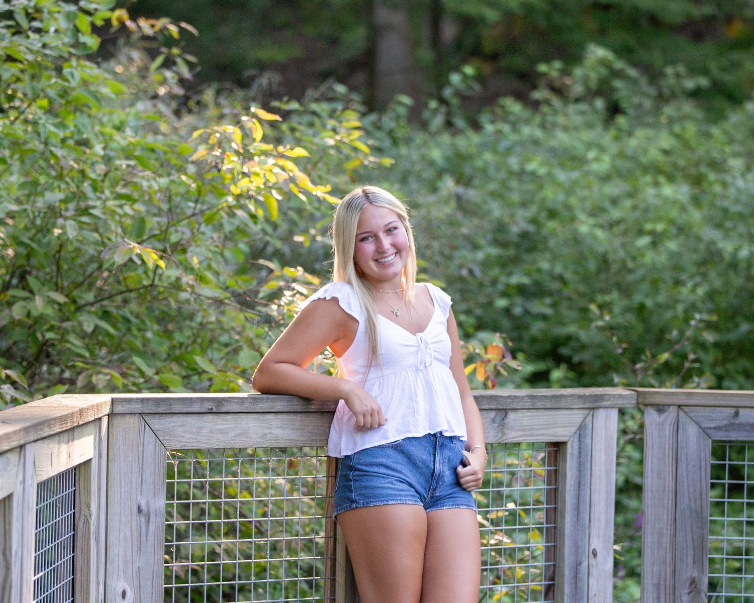 Young woman in a white shirt leaning, smiling, with trees and sunlight in the background.