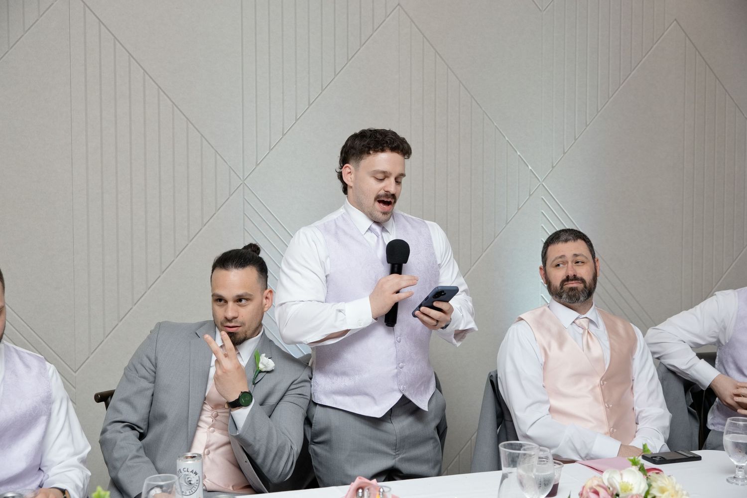 Groom giving a speech at wedding reception, holding microphone, surrounded by groomsmen in grey suits at head table.