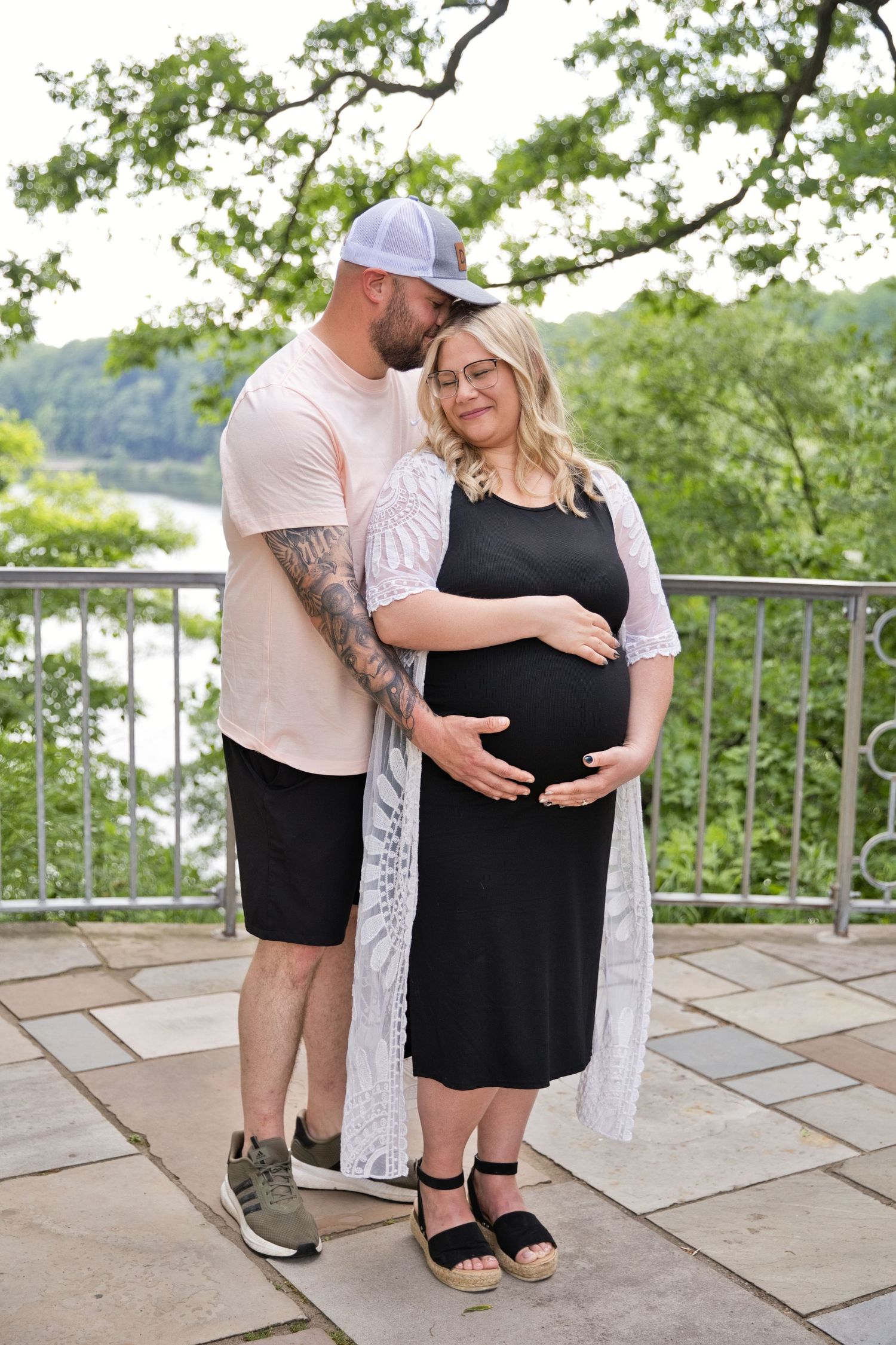 Couple poses outdoors during maternity photo shoot, man kisses womans head while she cradles baby bump.