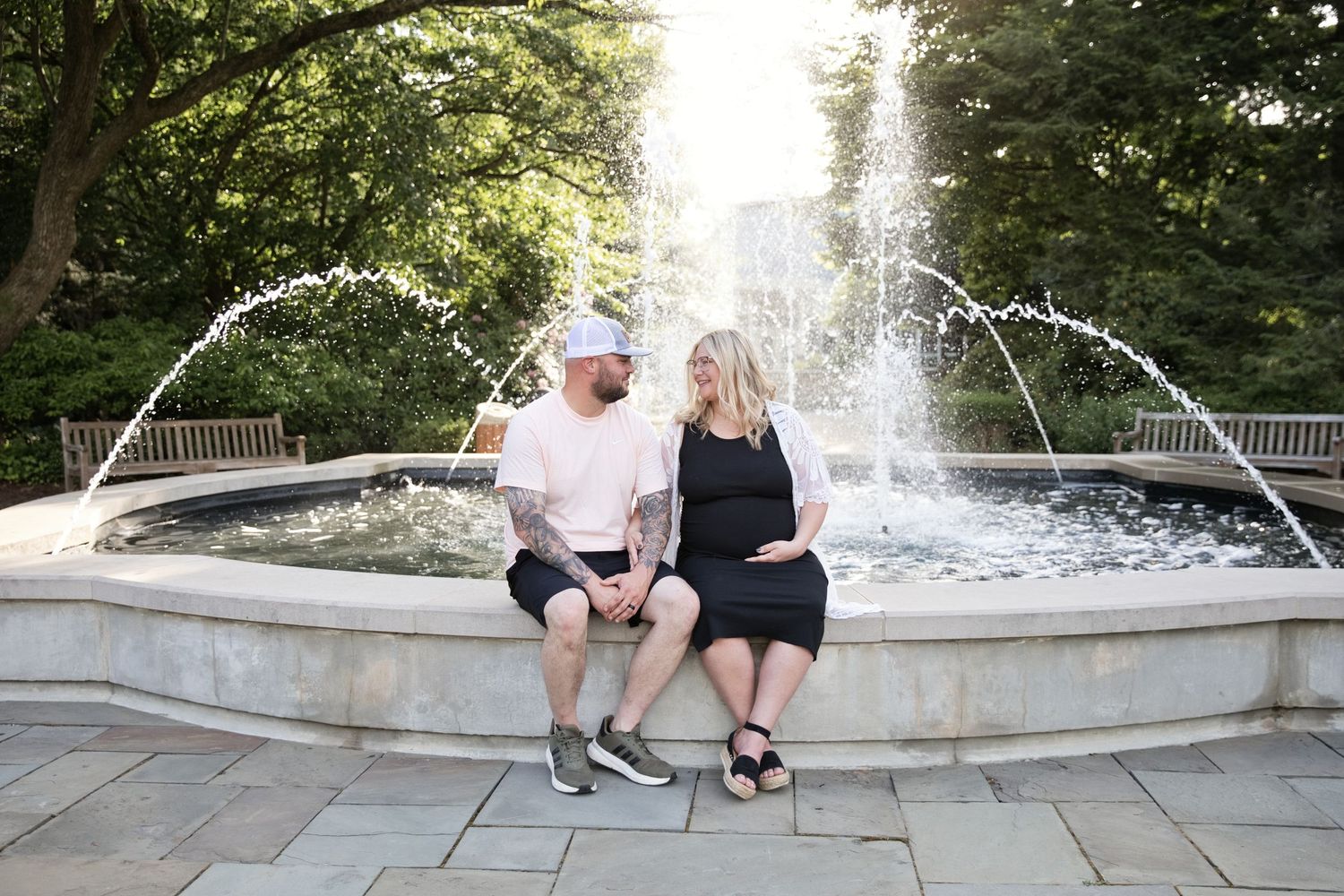 Pregnant couple sitting together by a garden fountain, man in pink shirt and white cap, woman in black dress.