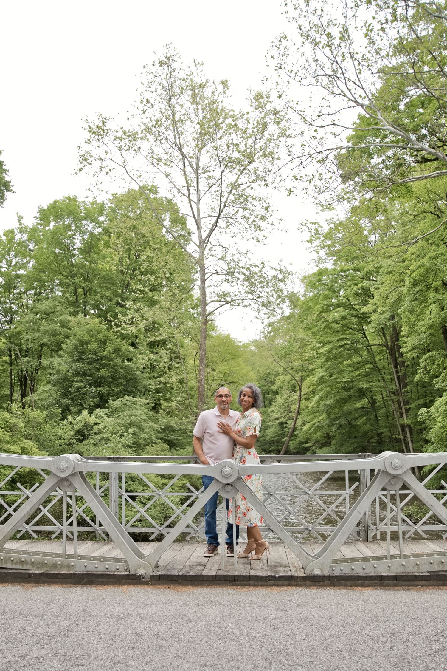 Couple embracing on a white ornate bridge surrounded by lush green trees in a park.