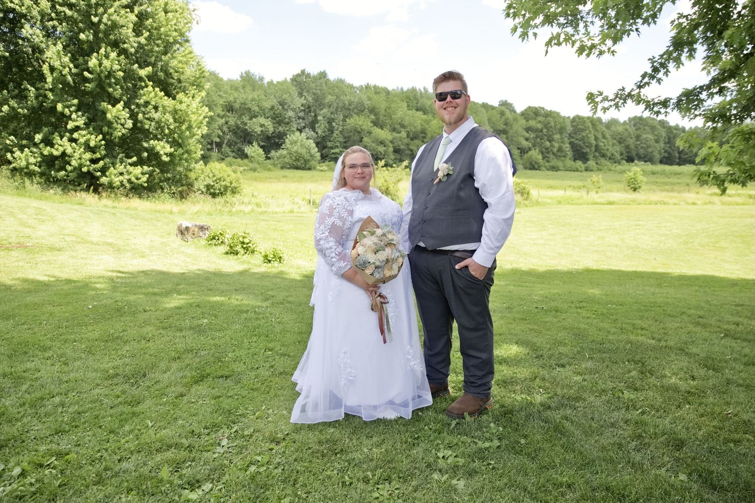 Bride and groom pose together on green grass at outdoor wedding ceremony, bride holding bouquet in white dress.
