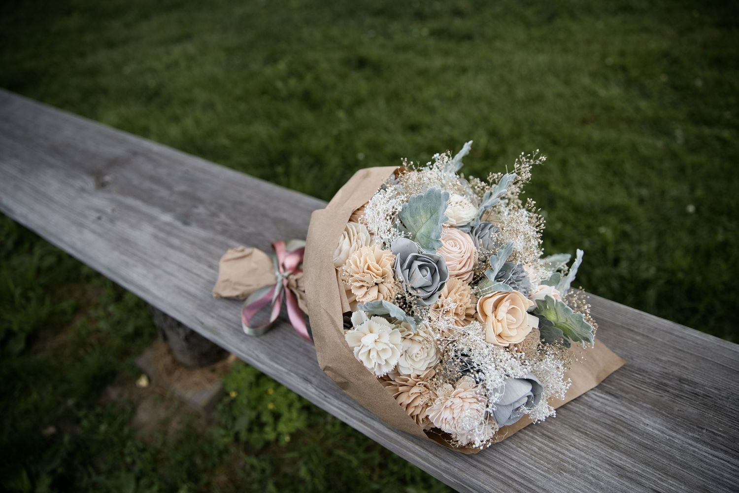 Rustic bridal bouquet with gray and blush sola wood flowers wrapped in kraft paper on a wooden bench.