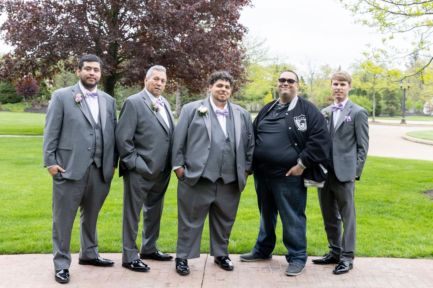Five men in gray suits with boutonnieres posing outdoors on a green lawn at a wedding celebration.