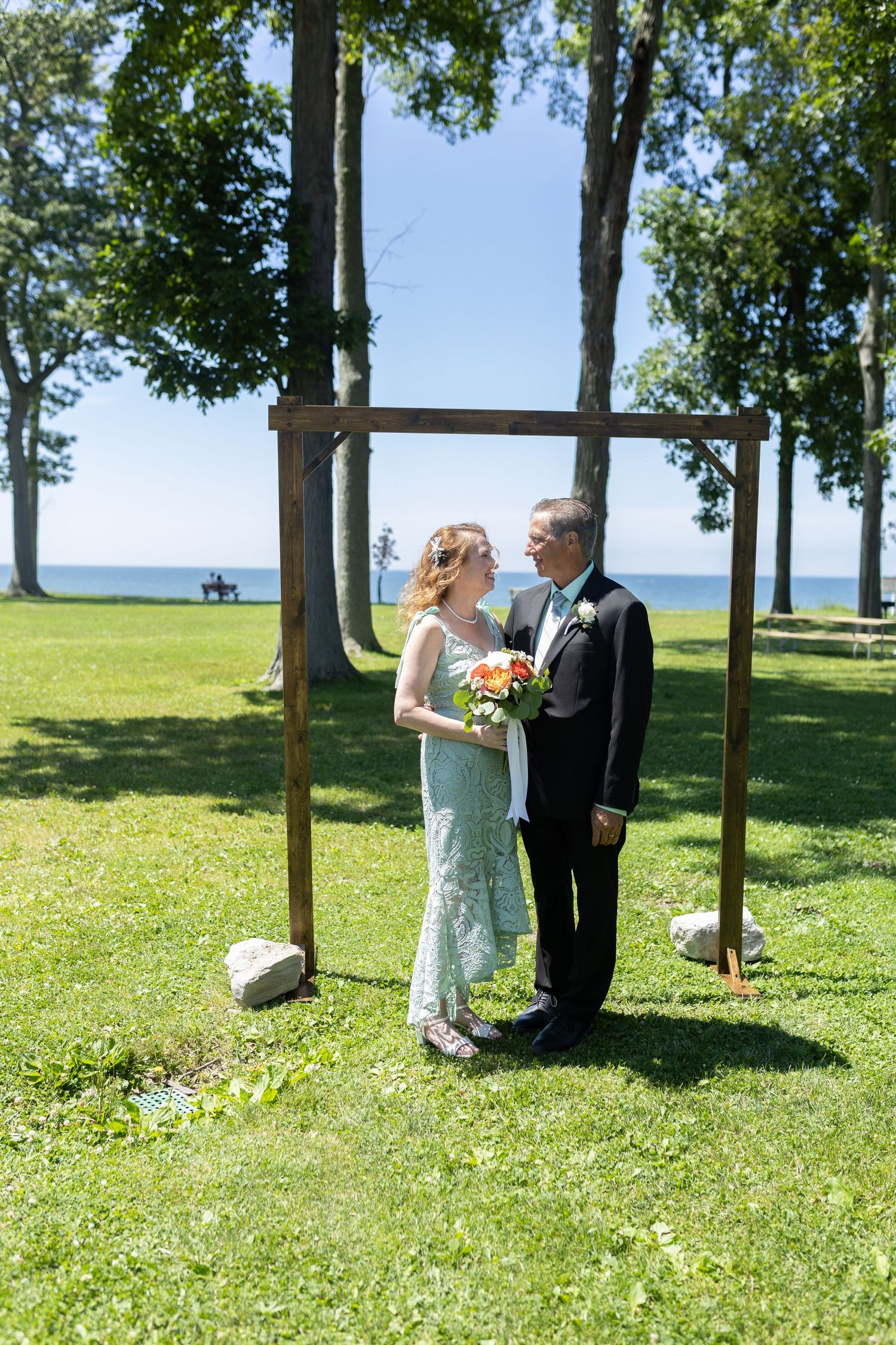 Outdoor wedding ceremony in a grassy field with trees and water view.