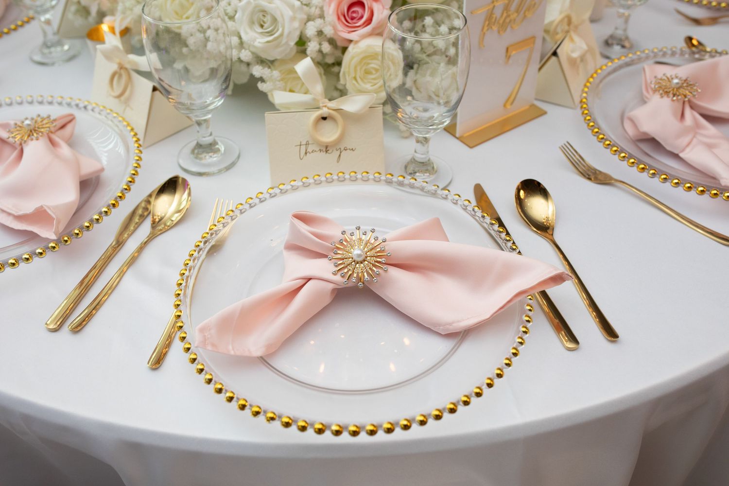 Elegant place setting with pink napkin, gold charger plate, and gold cutlery on white tablecloth.