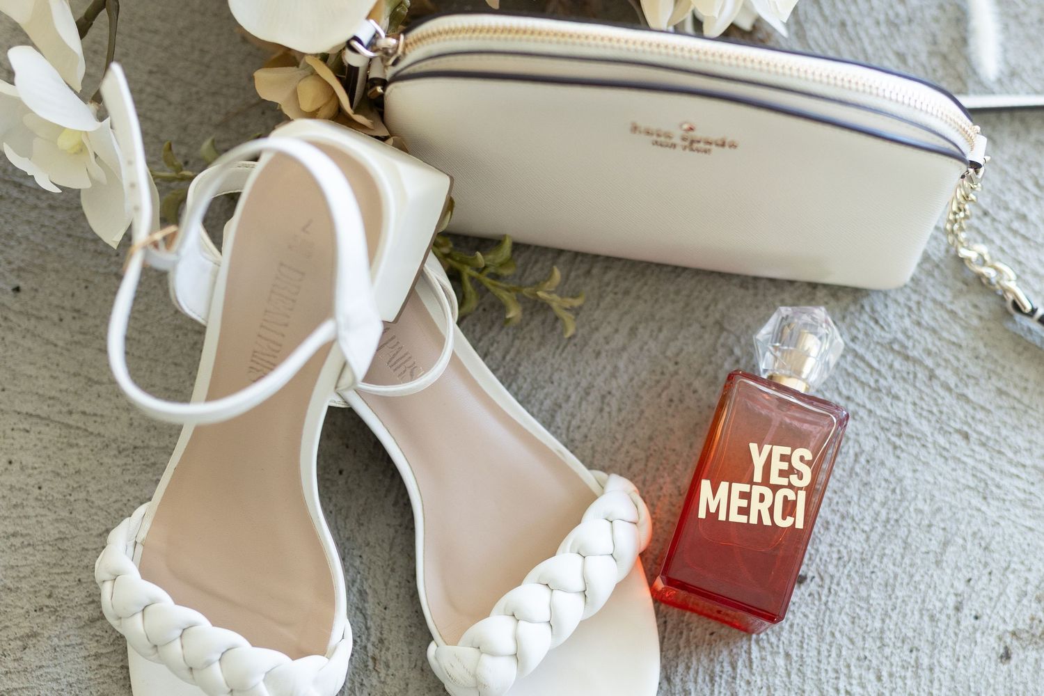 White bridal shoes and clutch displayed with a small red perfume bottle labeled Yes Merci.