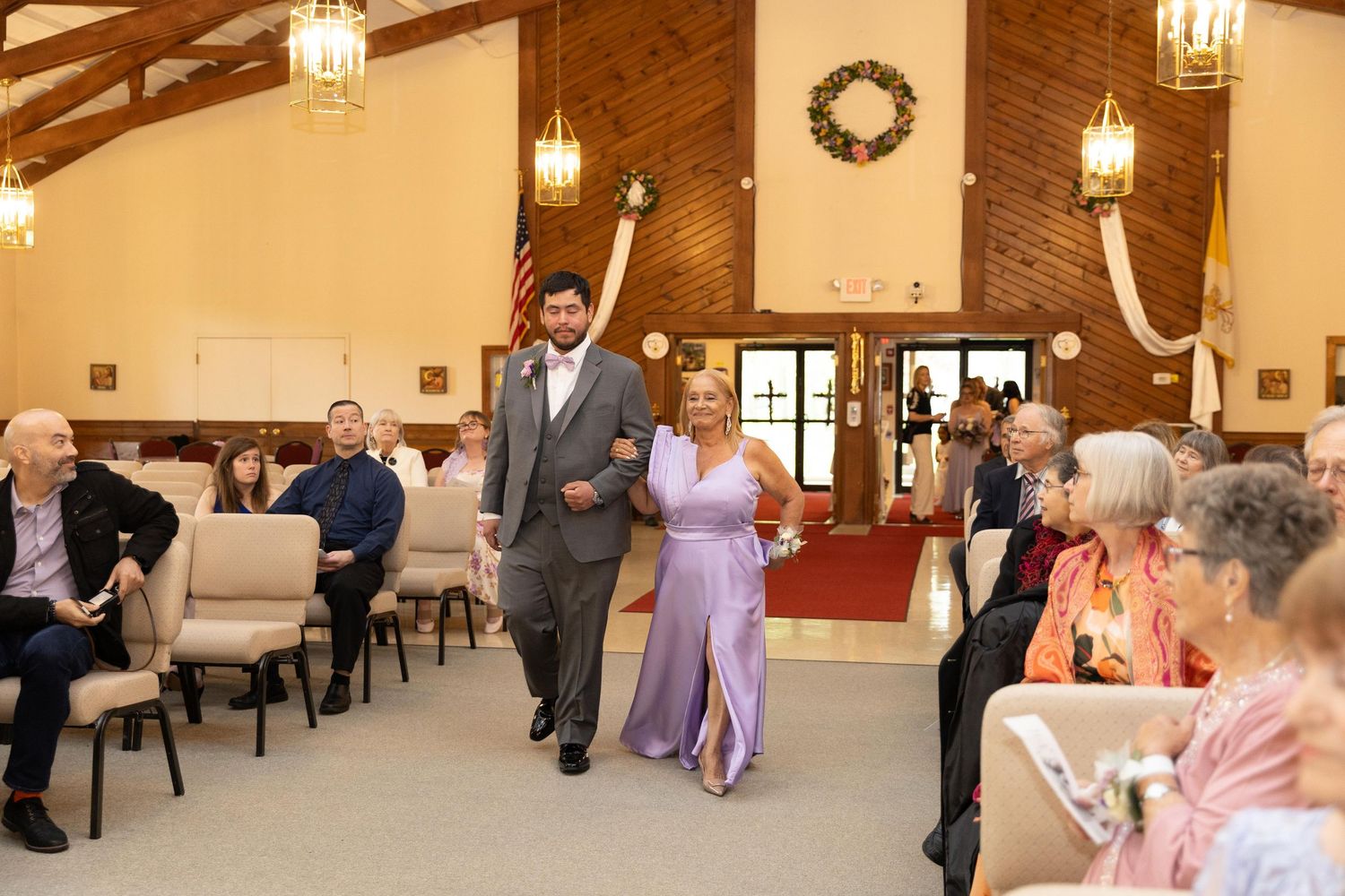 Couple walks down wedding aisle in elegant venue, man in gray suit, woman in lavender gown, guests seated nearby.